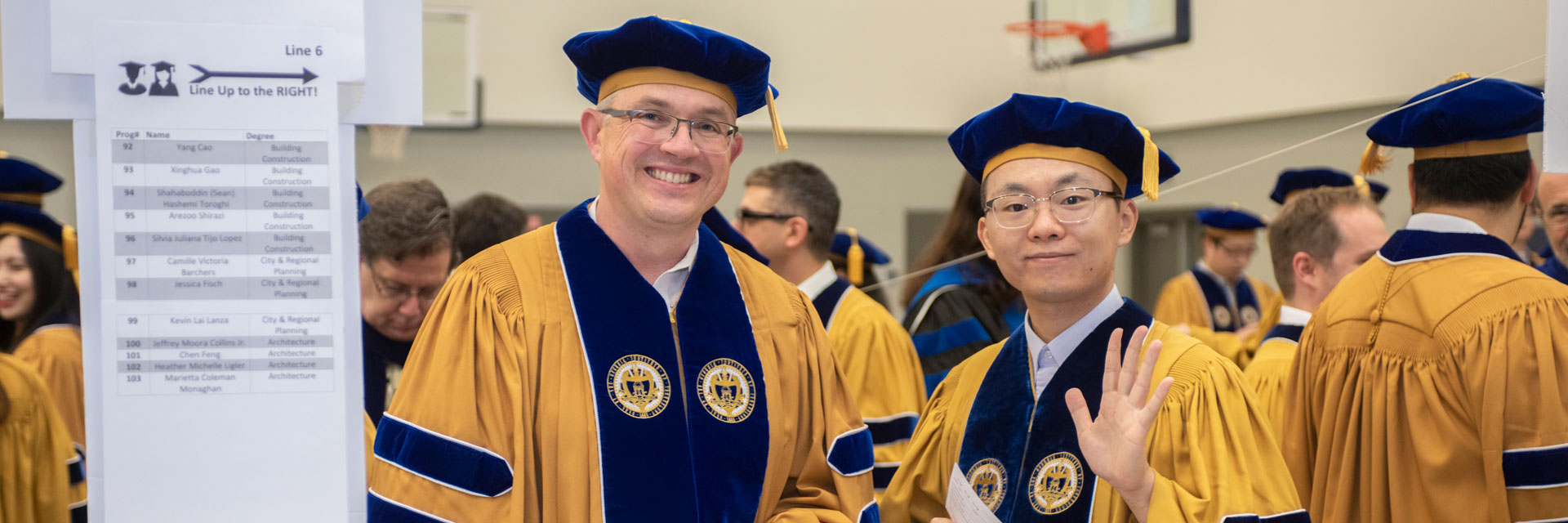 Two Ph.D. graduates standing in line at commencement ceremony. One waving to the camera. 