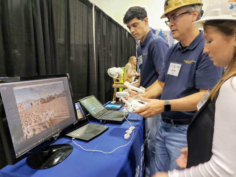 A male professor with a remote in his hand looks at a drone simulation on a monitor with a male and female student standing by his side. 