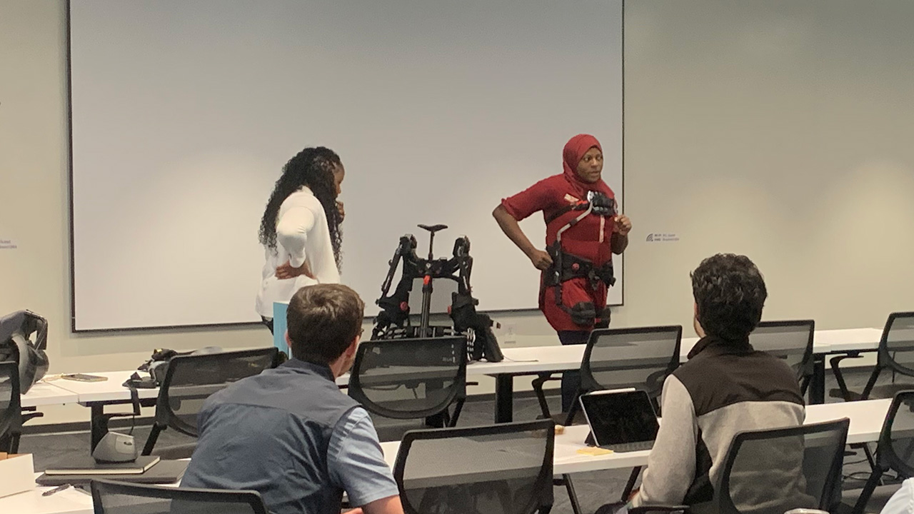 Two people demonstrating exoskeleton wear and use in front of a conference audience.